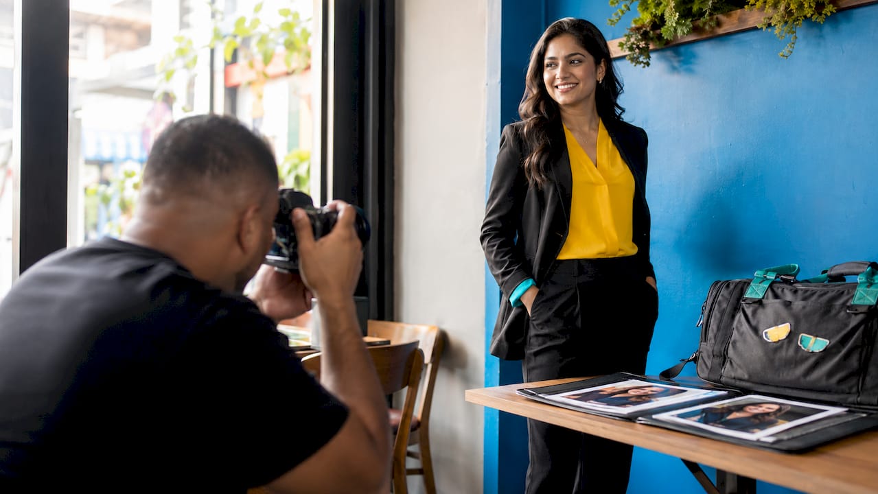 Photographer taking a portrait of a woman standing by a blue wall with a table of camera gear nearby