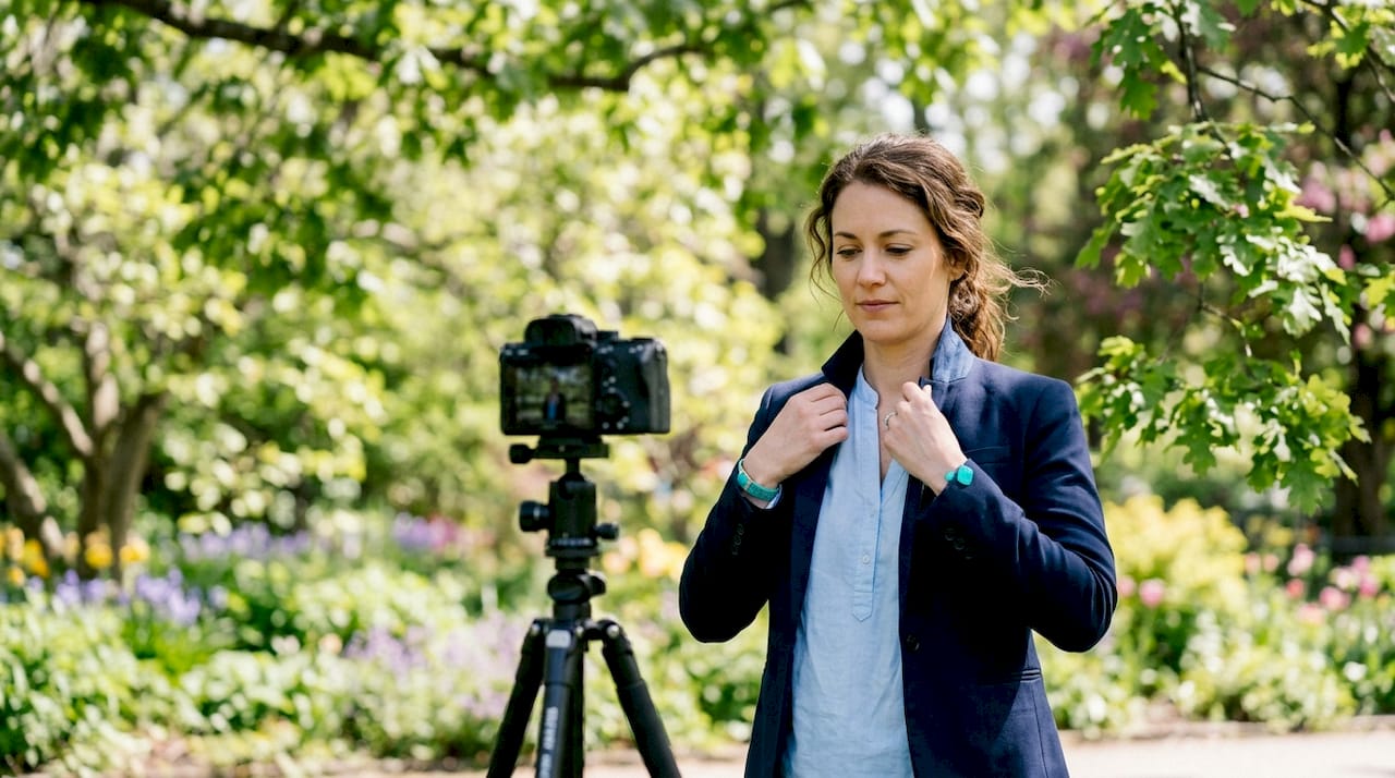 Woman adjusting her collar beside a camera on a tripod in a leafy garden