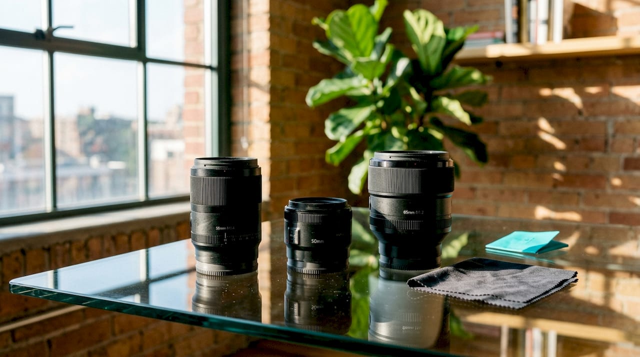 Camera lenses on a glass table by a sunlit window, with a plant and brick wall in the background