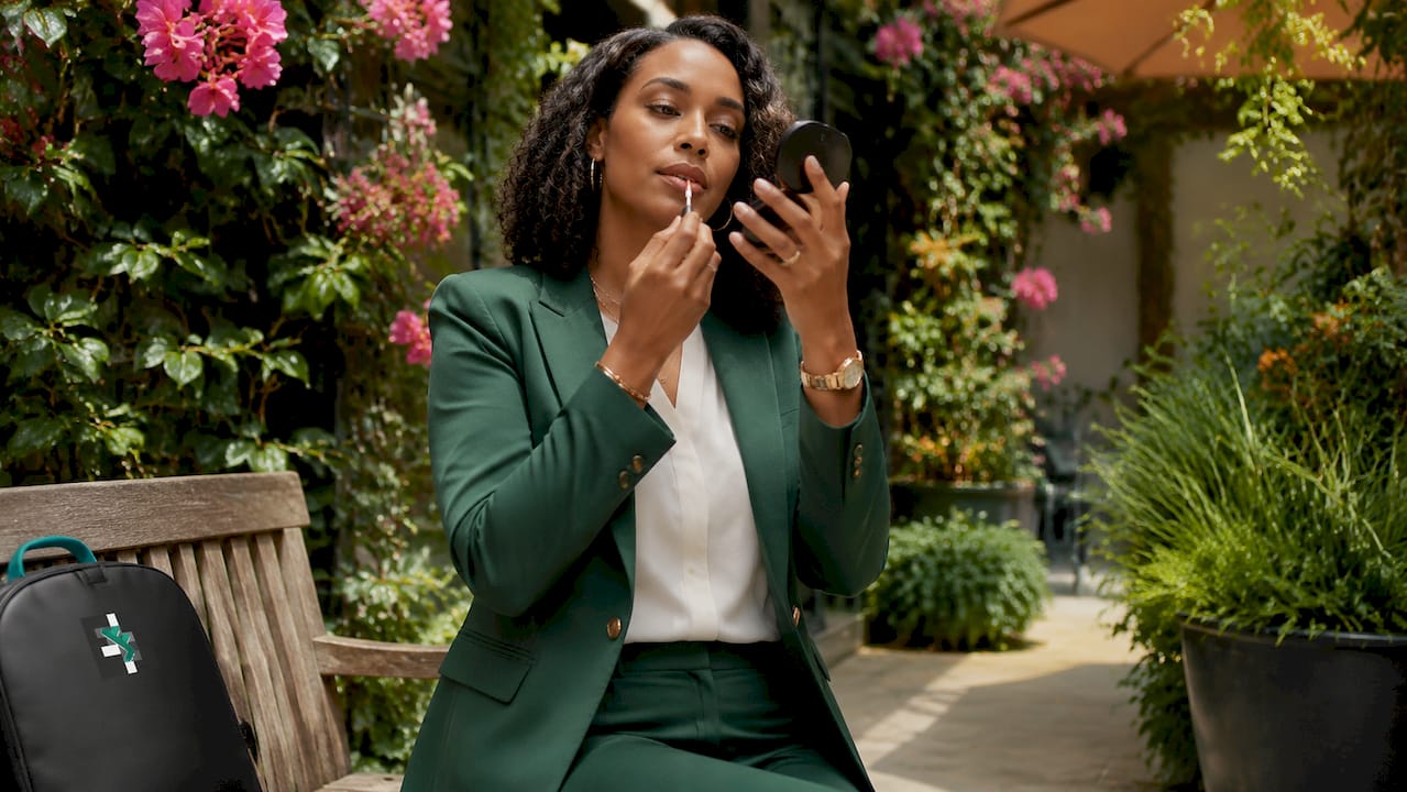 Woman in a green suit applying lipstick while sitting in a garden patio with flowers and plants.