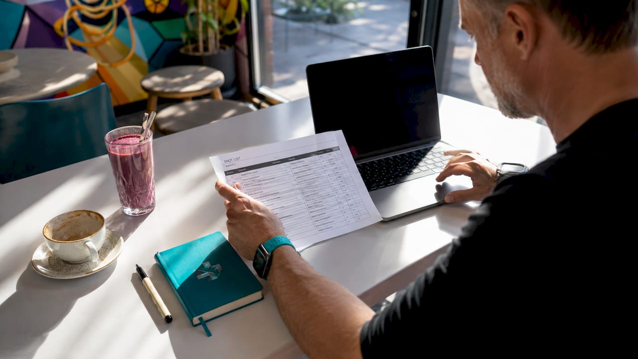 Person reviewing a document at a desk beside a laptop, coffee, and notebook by a window.