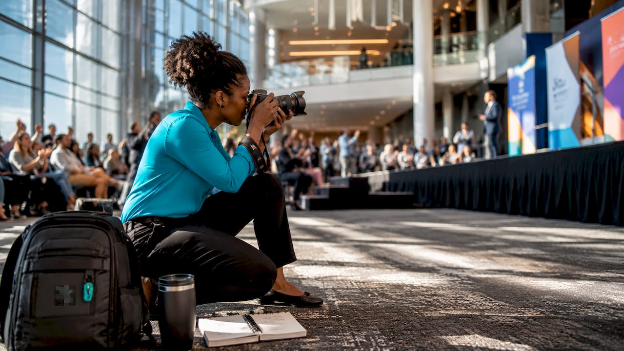 Person sitting on the floor at a busy conference, taking notes with a camera bag beside them.