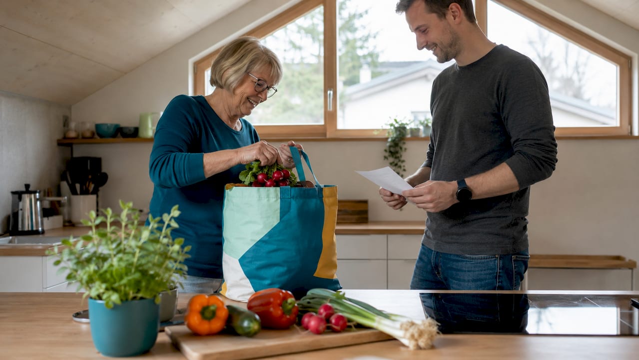 Eine Seniorin wird beim Ausräumen der Einkäufe in ihrer Küche von einem Helfer unterstützt.