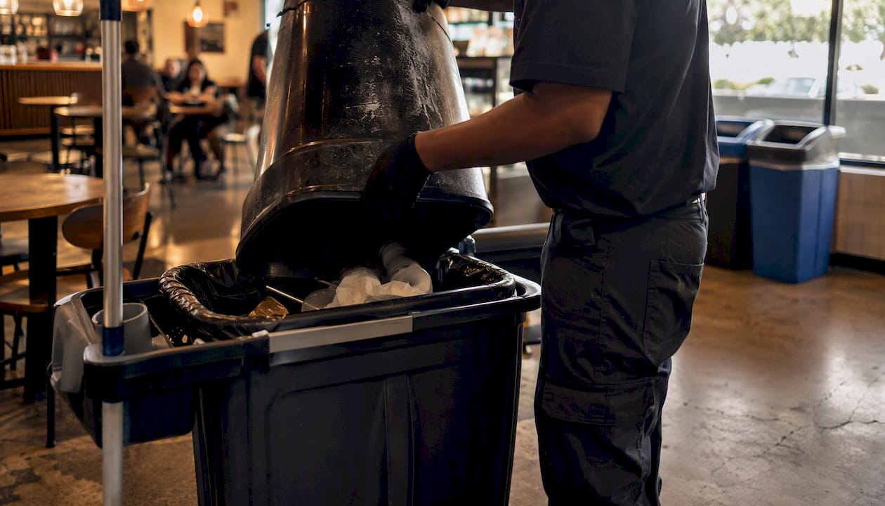 Why Trash Management Is Part of Janitorial Services 1 Janitor emptying trash bin in office corridor
