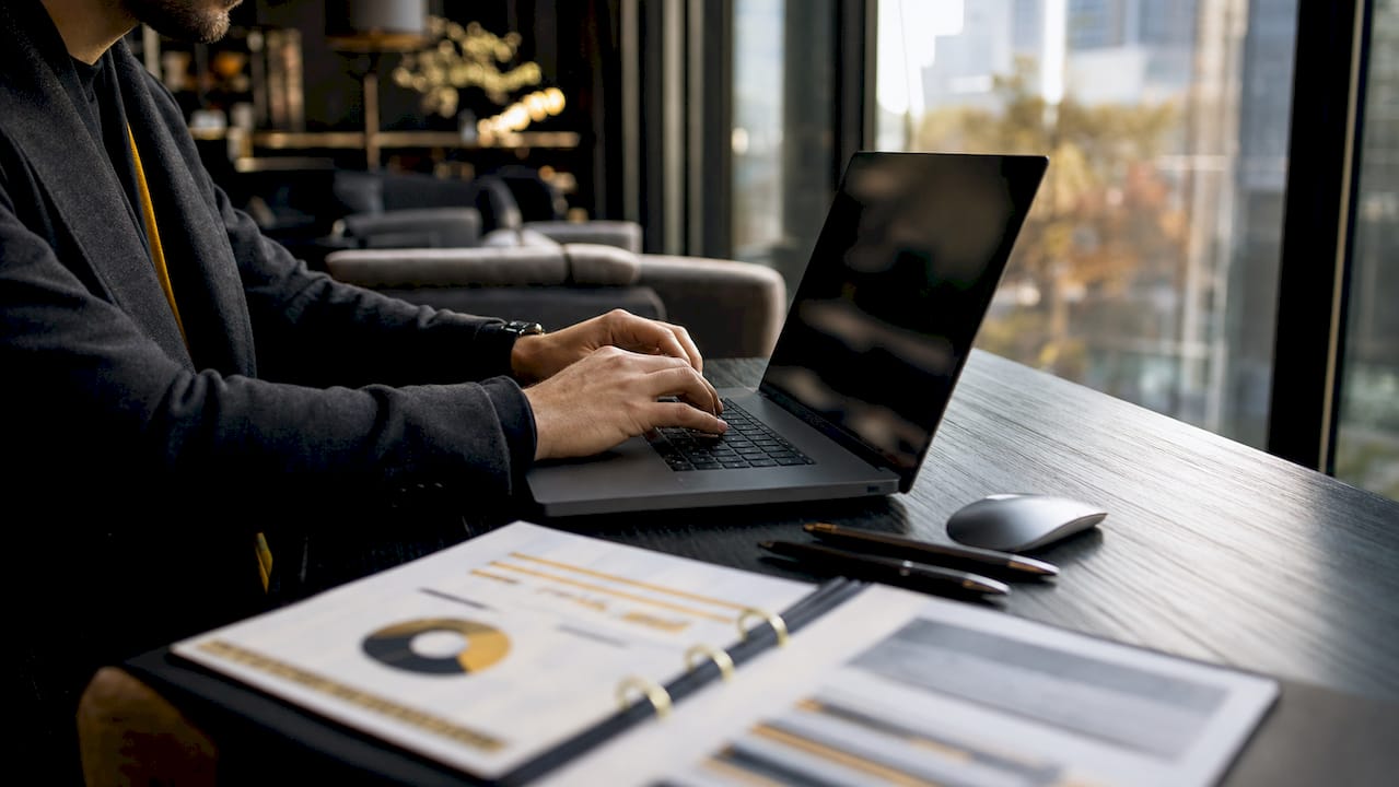 Hands typing on laptop with financial reports on table