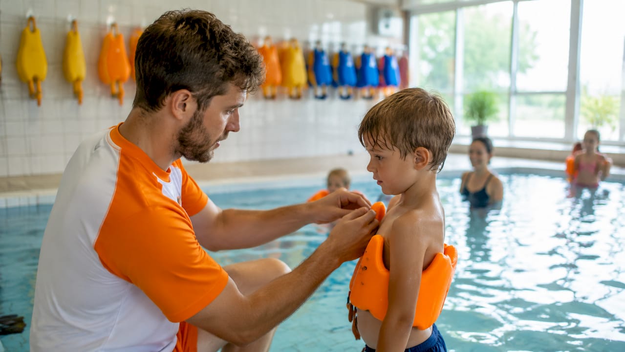 Ein Schwimmlehrer unterstützt ein Kind mit Schwimmflügeln im Wasser.