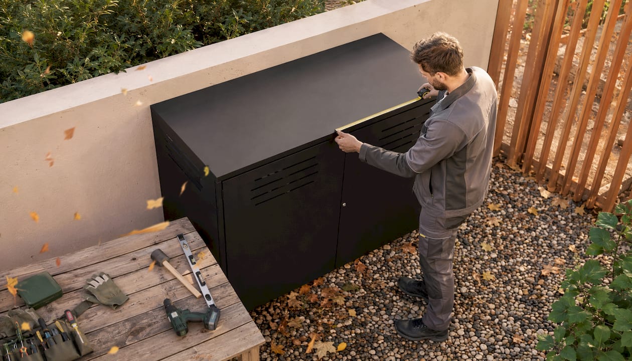 Ein Handwerker nimmt vor dem Haus Maß für eine Mülltonnenbox aus Aluminium.
