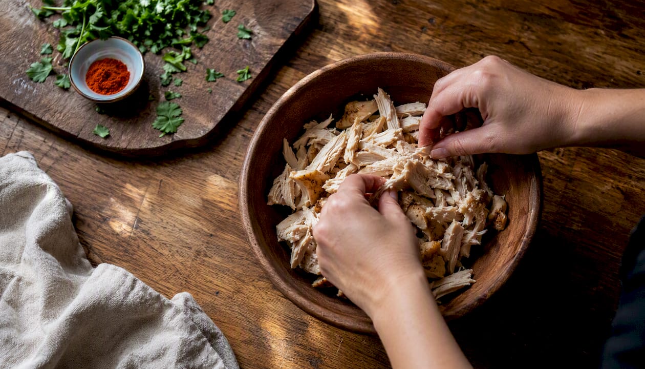 Hands shredding cooked chicken over bowl on butcher block