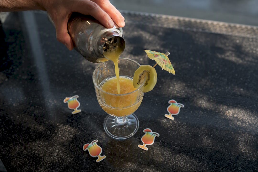 Hand pouring tropical cocktail with emoji cutouts nearby
