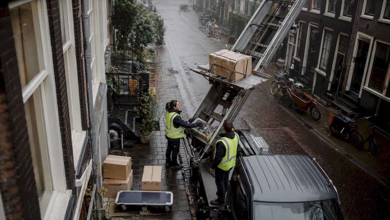 In een smalle straat in Amsterdam zijn verhuizers druk in de weer met het bedienen van een verhuislift.