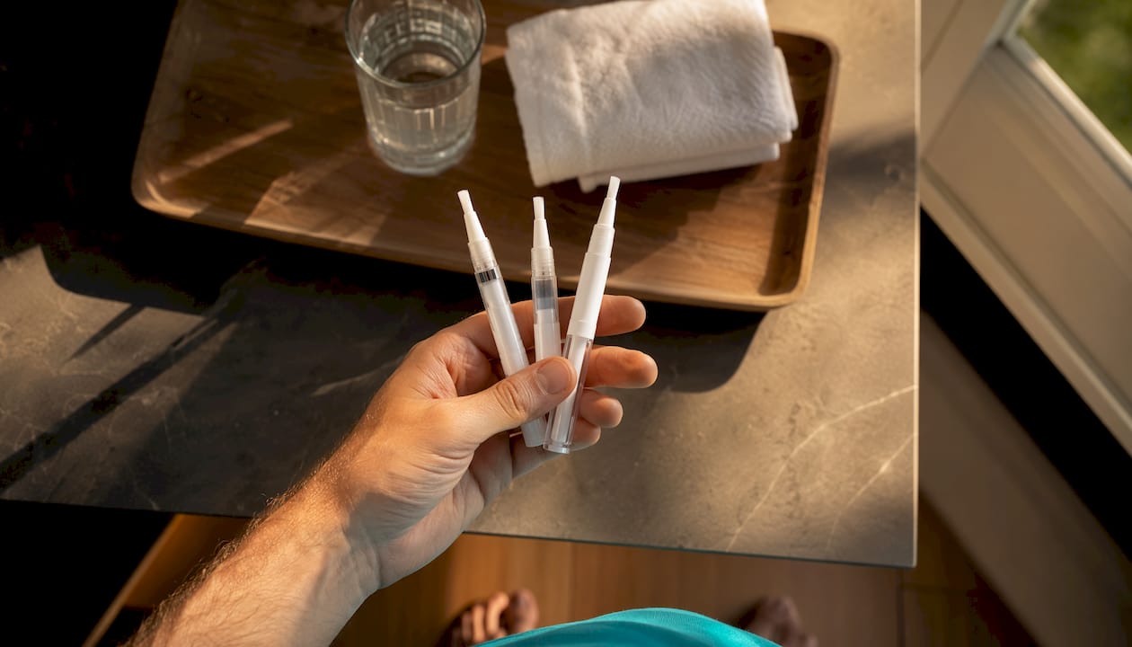 Una mano che tiene diverse penne sbiancanti appoggiate sul lavandino del bagno.
