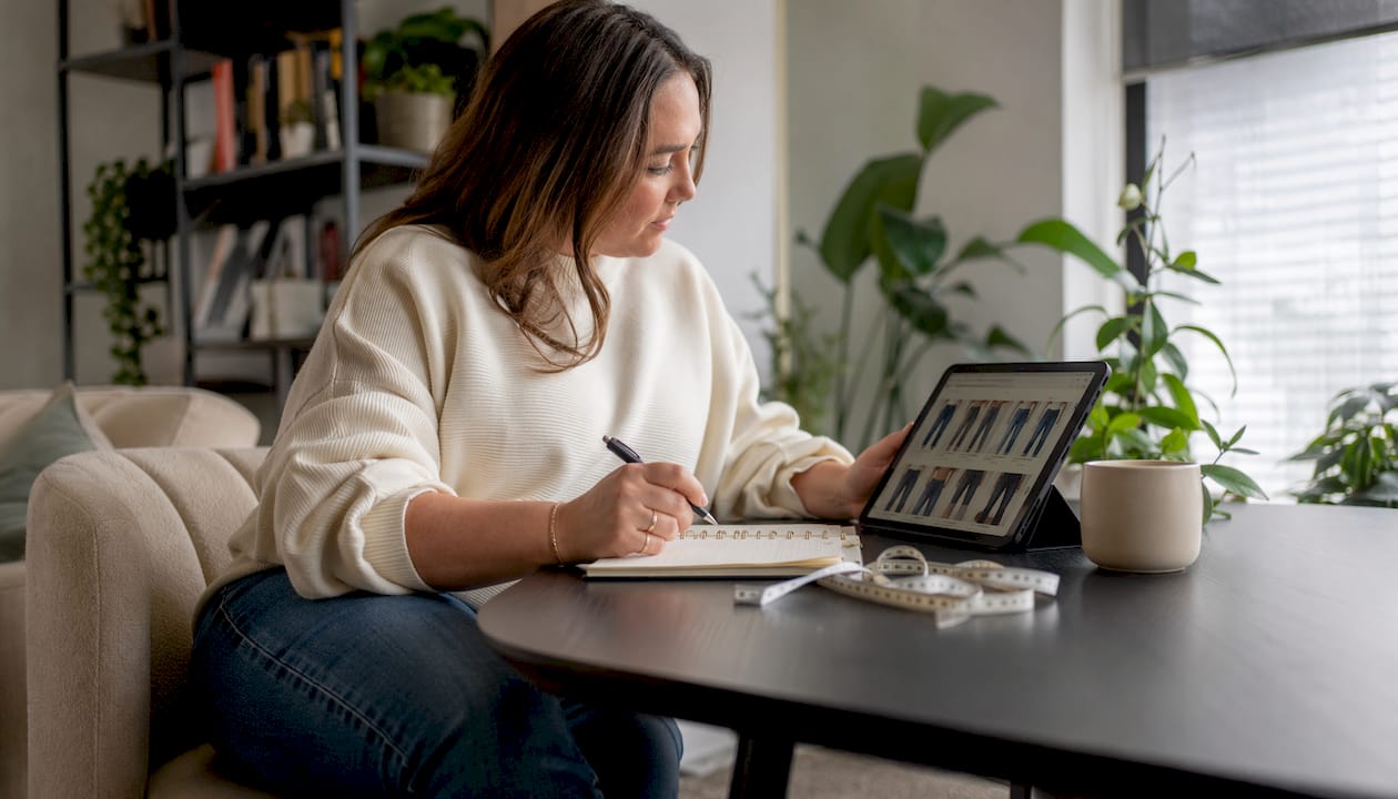 Een vrouw met een klein postuur en een maatje meer shopt online naar spijkerbroeken terwijl ze aan de eettafel zit.