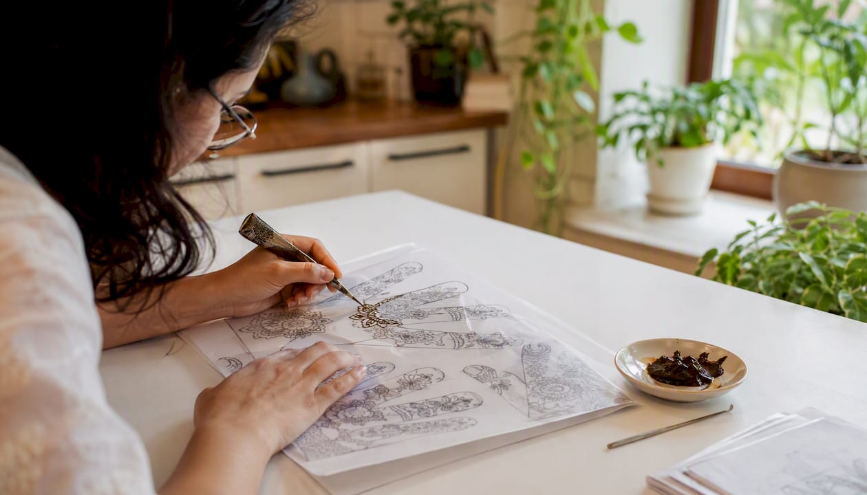 Hands tracing henna motif with mehndi cone over plastic overlay