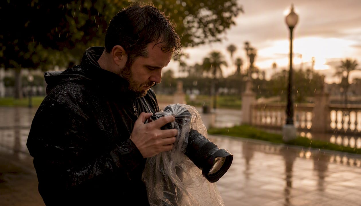 Un fotógrafo ajusta su cámara bajo la lluvia, buscando el encuadre perfecto pese al mal tiempo.