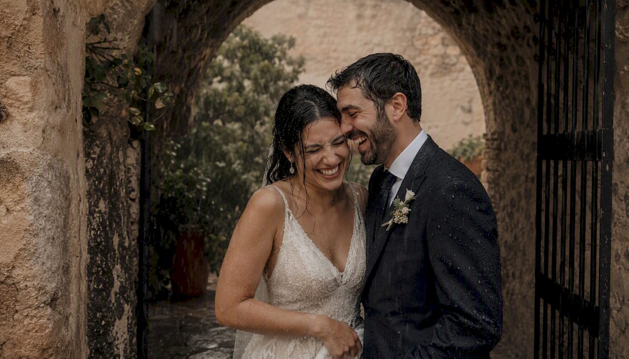 Una pareja de recién casados se ríe a carcajadas bajo un arco de lluvia.