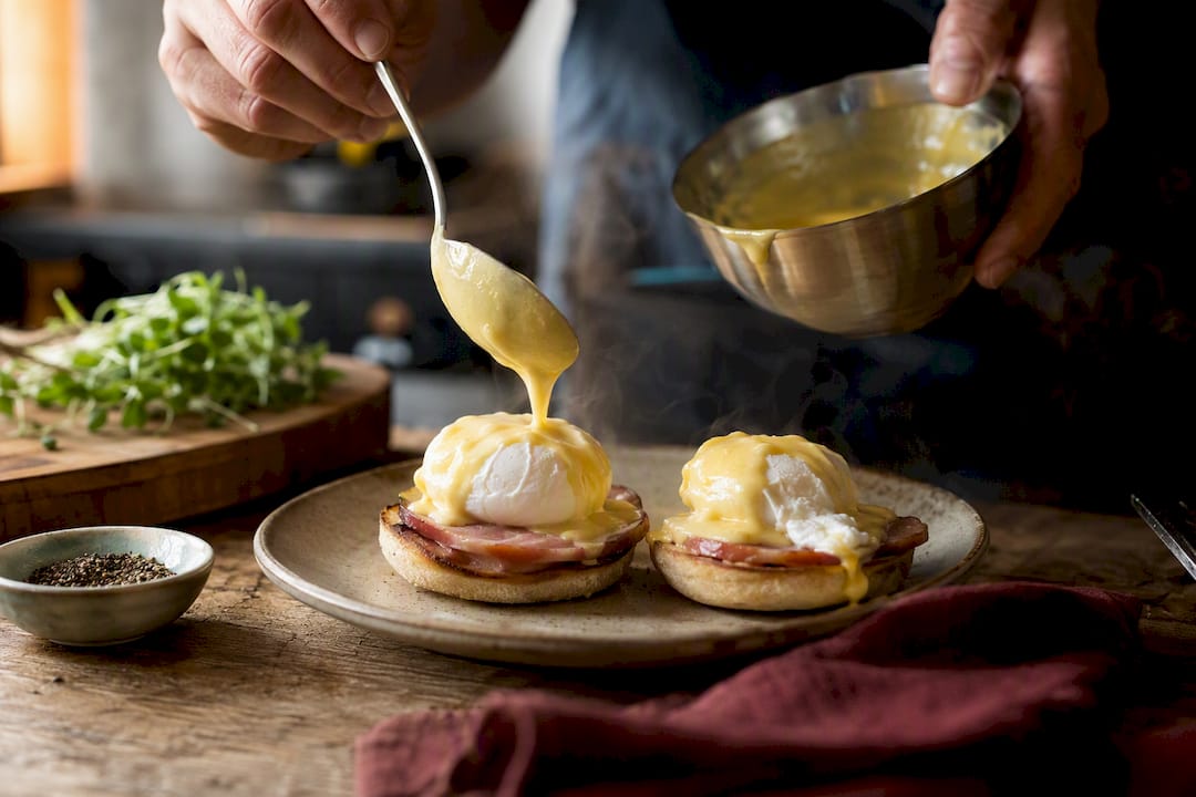 Hands spooning hollandaise on eggs benedict