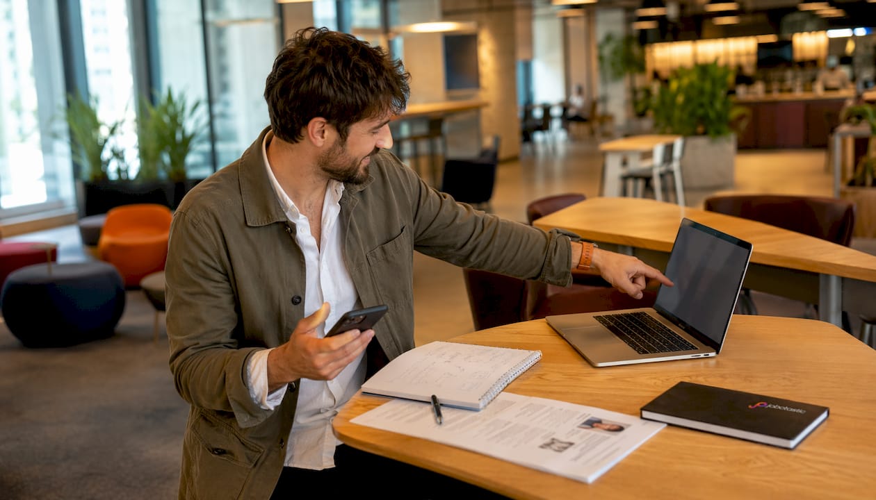 Ein Mann präsentiert im Büro sein Smartphone und seinen Laptop.