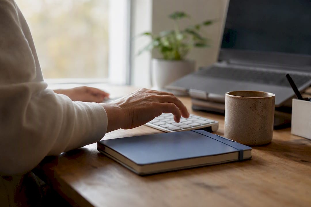 Someone typing notes on their laptop, with a steaming mug next to the keyboard.