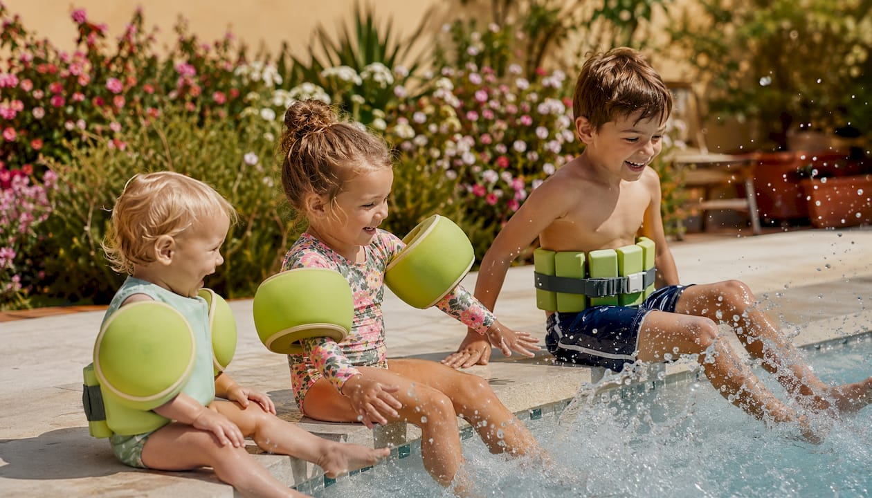 Am Beckenrand planschen Kinder fröhlich mit Schwimmflügeln, Poolnudeln und anderen Auftriebshilfen im Wasser.