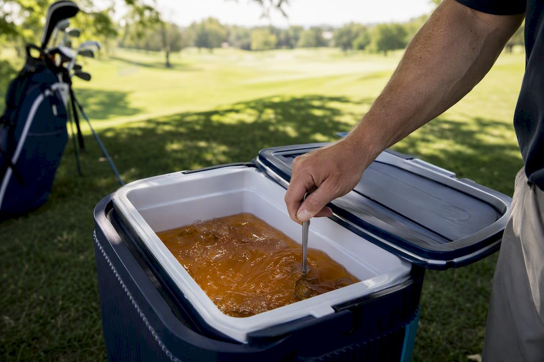 Hand stirring iced tea batch in cooler by golf bag