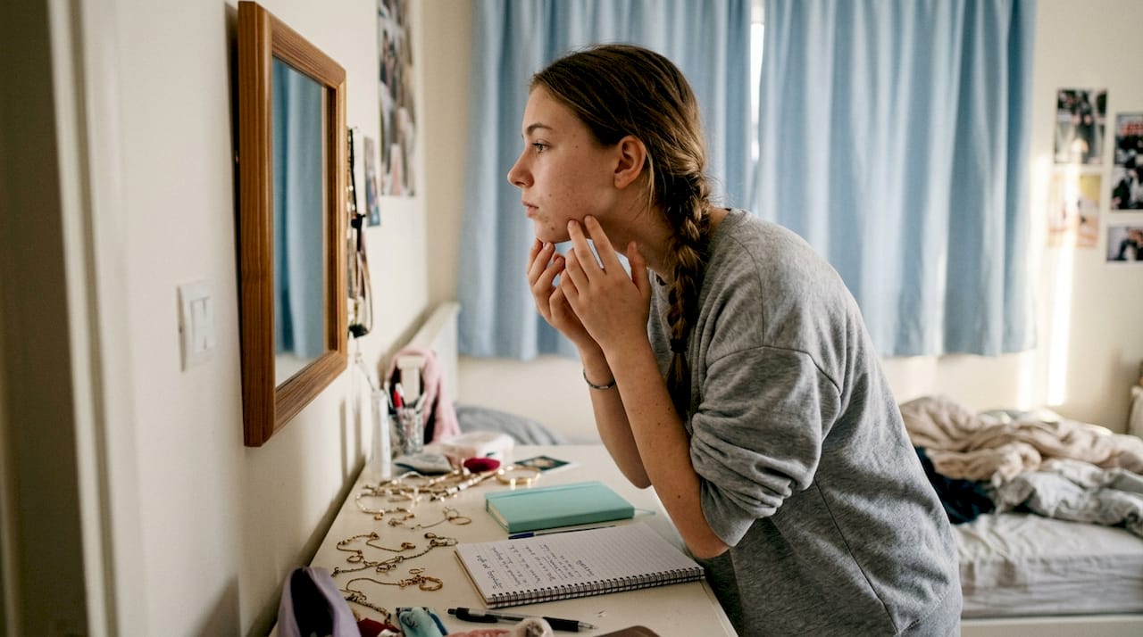 Acne prone δέρμα: Τι σημαίνει και πώς να το φροντίσετε 2 Έφηβη κοπέλα παρατηρεί προσεκτικά το σαγόνι της στον καθρέφτη για σημάδια ή σπυράκια.