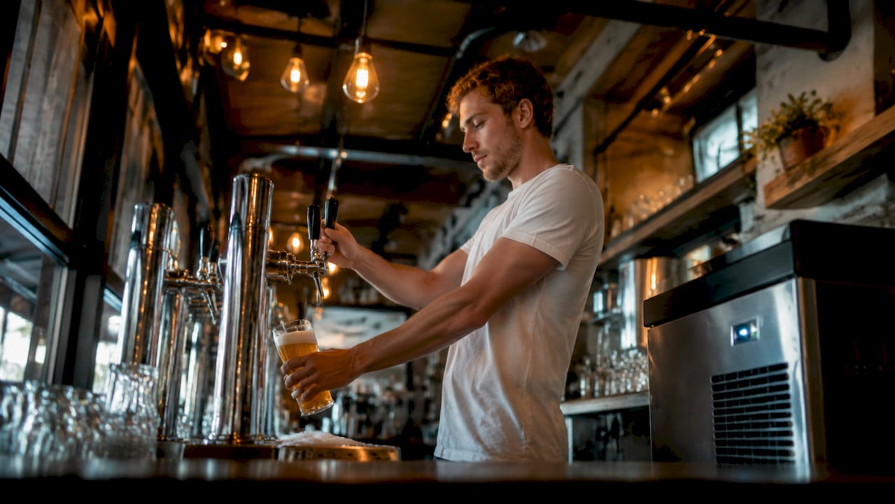 Achter de bar staat de barman druk biertjes te tappen voor de volle zaak.
