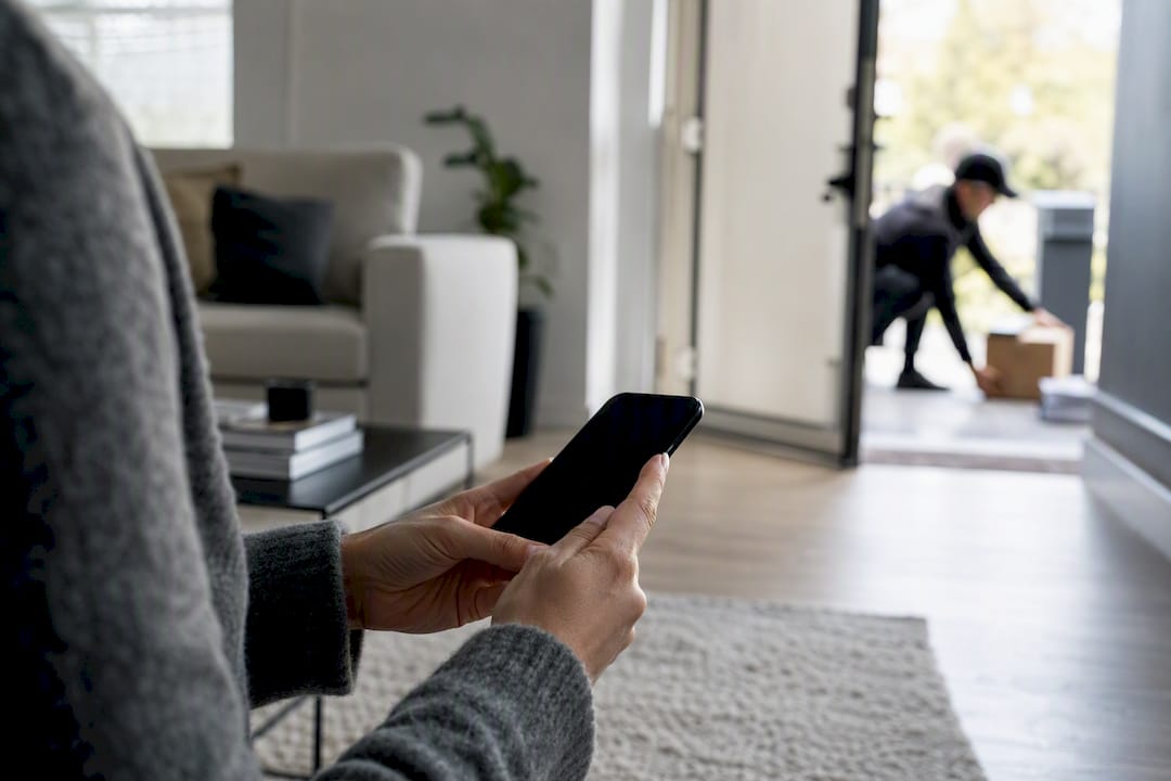 Handen die een telefoon vasthouden bij een bezorgd pakketje op de stoep