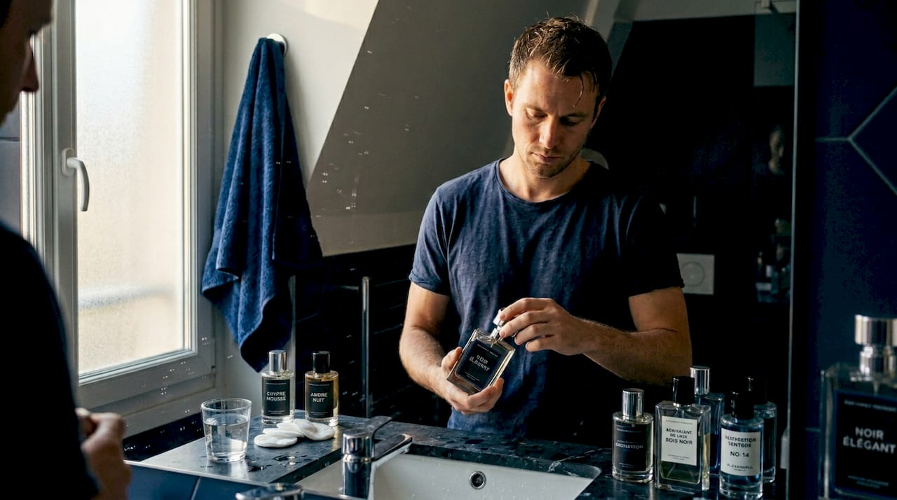 Un homme se tient devant son lavabo, examinant attentivement deux flacons de parfum identiques pour tenter de déceler la moindre différence.