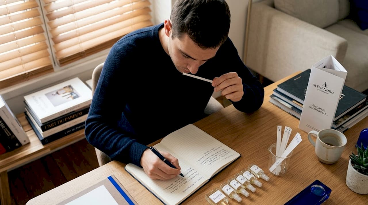 Assis à son bureau en bois, un homme prend le temps d’examiner différents parfums, concentré sur l’art délicat de la découverte olfactive.