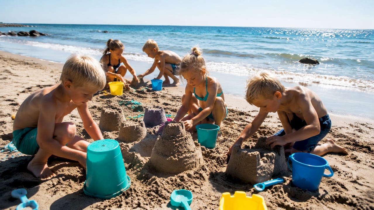 Små børnehænder graver og bygger med spande og skovle i sandet på stranden.
