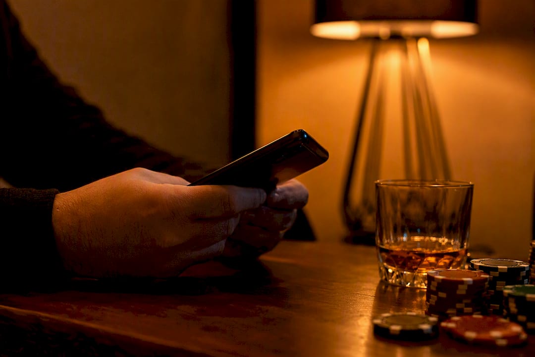 Manos sosteniendo un smartphone junto a fichas y whisky
