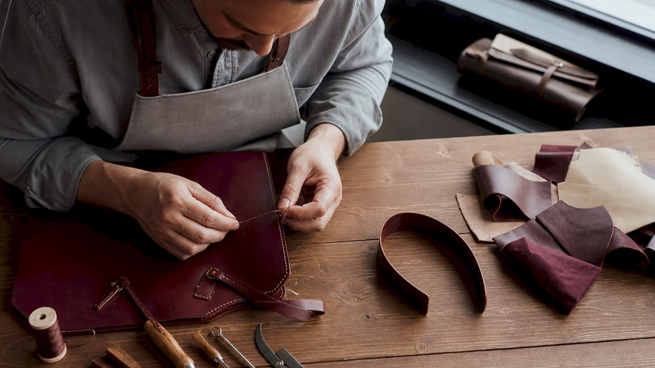 Detailopname van een vakman die met de hand een luxe leren tas stikt.