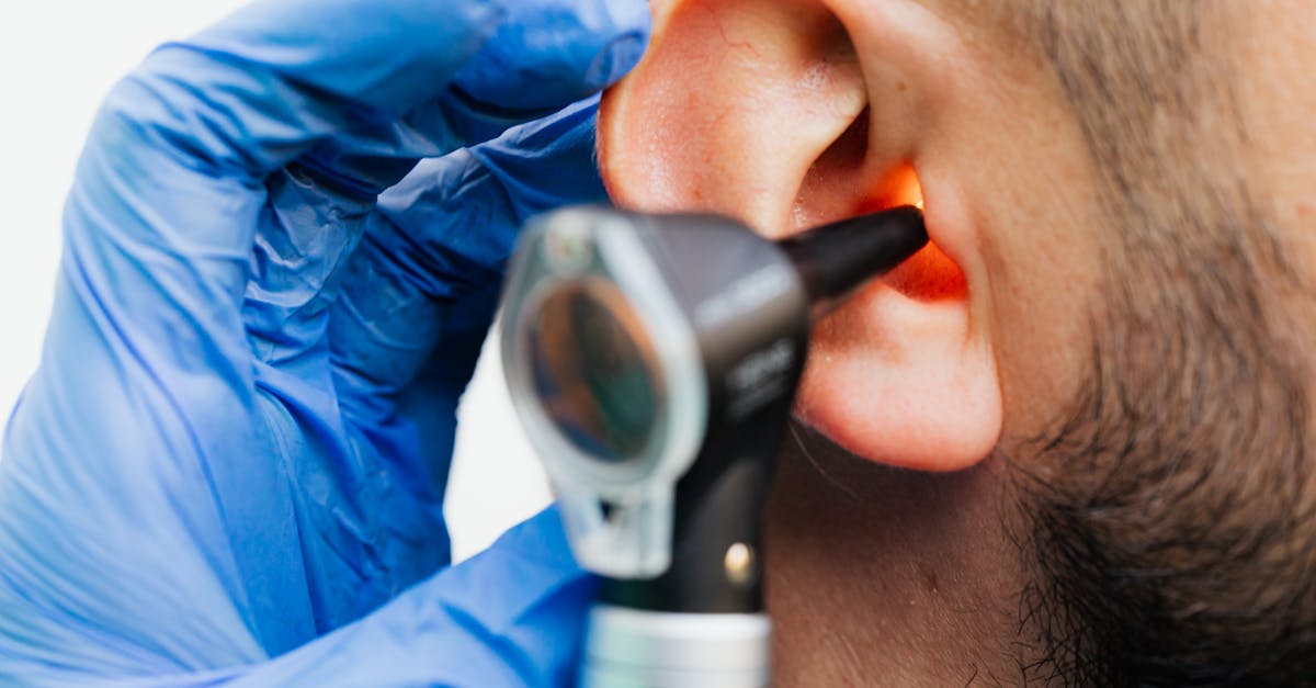 Otoscope inspection of an ear by a person in blue gloves. Bright light illuminates the ear canal. Close-up, clinical setting.