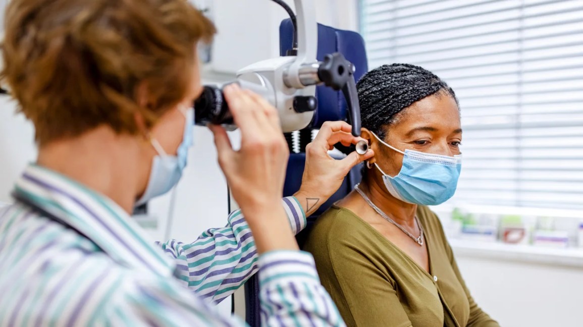 A doctor examines a patient with an eye device in a medical office. Both wear masks. The patient looks calm. Blinds and shelves in background.