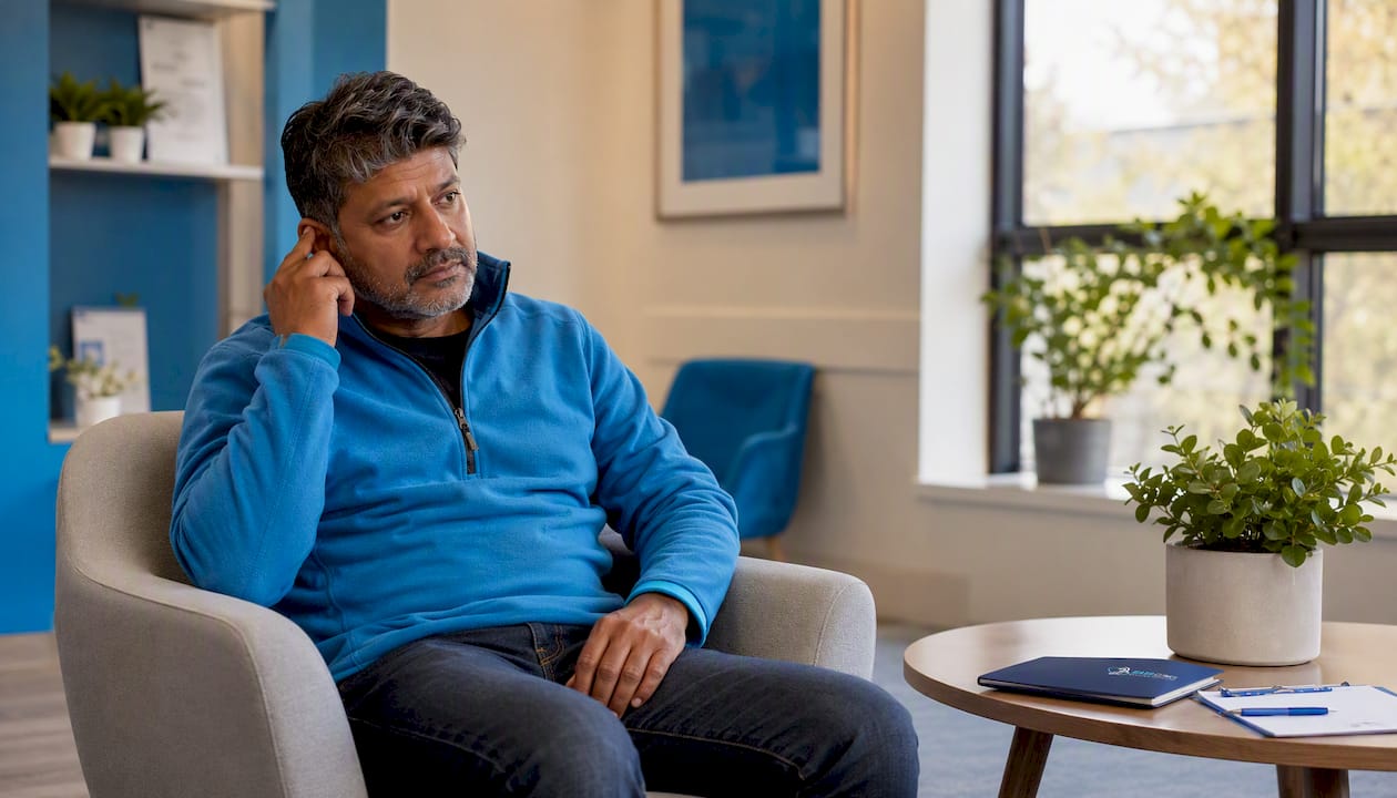 Man seated in clinic waiting area touching ear thoughtfully